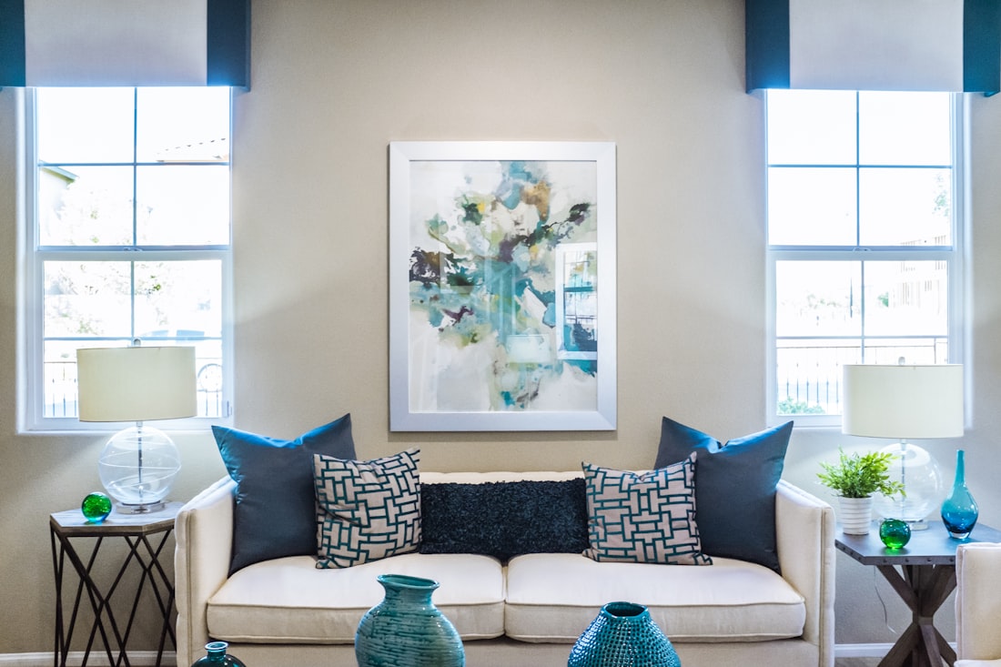 White sofa with teal and navy cushions under an abstract painting, with a lamp on a side table at each window