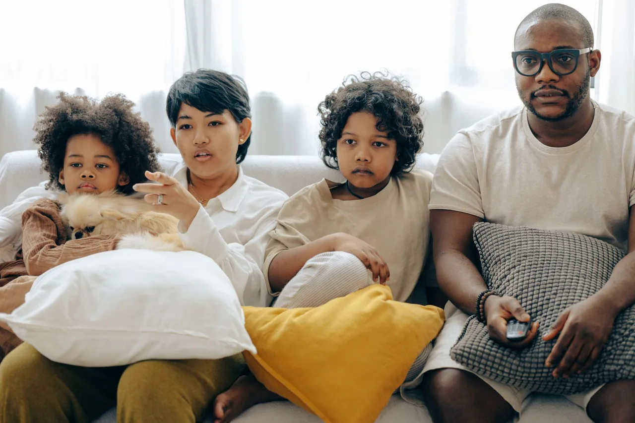 Two adults and two children on a white sofa watching TV, the man in glasses holding the remote