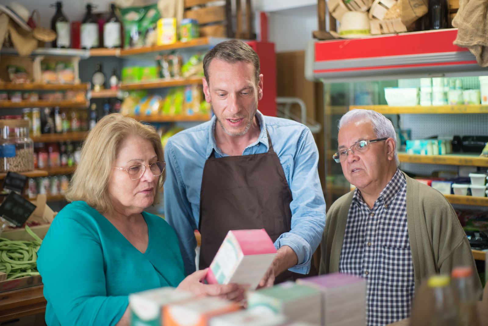 A shop assistant in a brown apron shows a product box to an older couple
