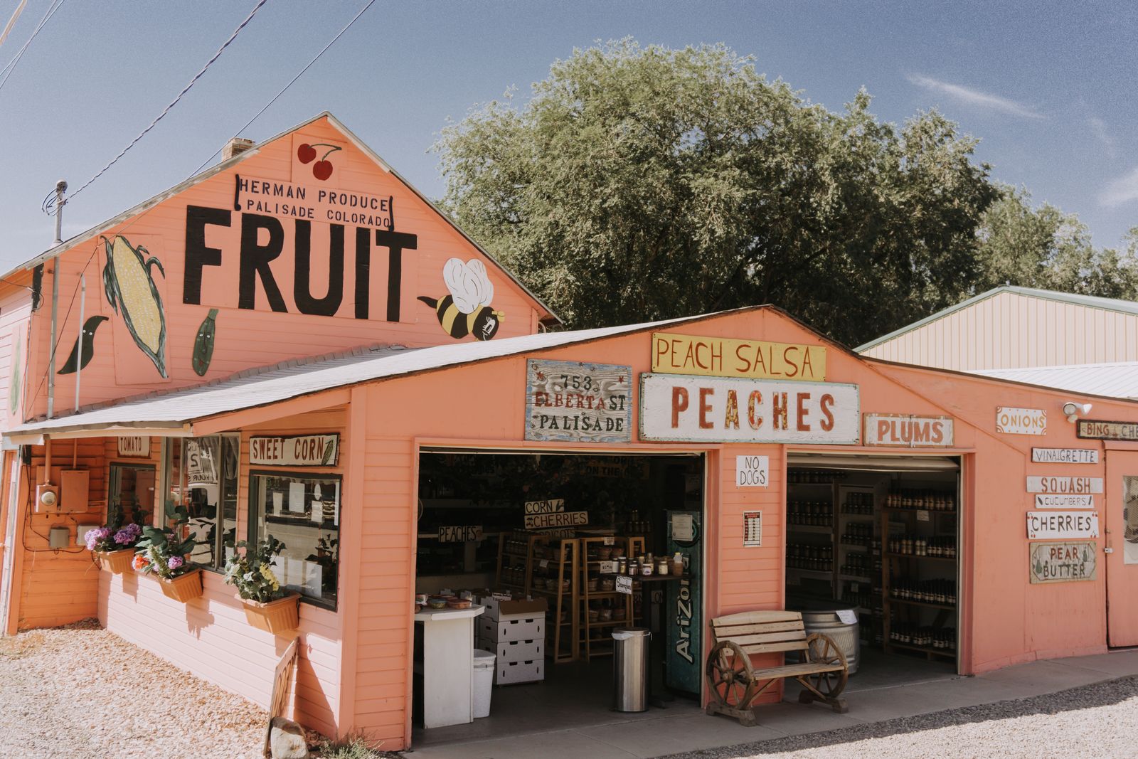 Peach-painted roadside produce stand with hand-lettered signs for peaches, plums and sweet corn