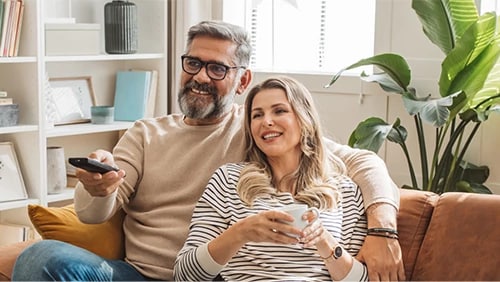 A man with glasses aims a remote while the woman beside him holds a mug on a tan sofa