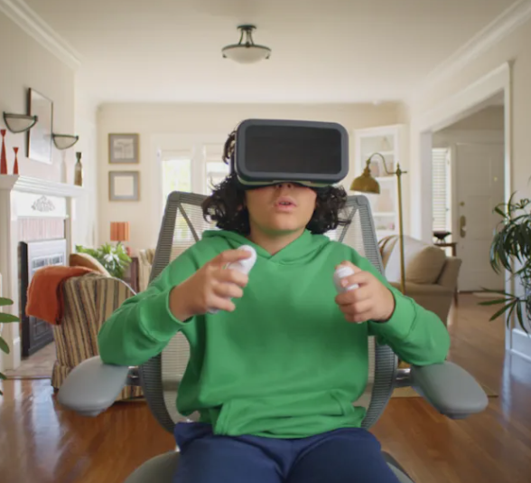 A child in a green hoodie sits in a mesh office chair playing with a VR headset and two controllers