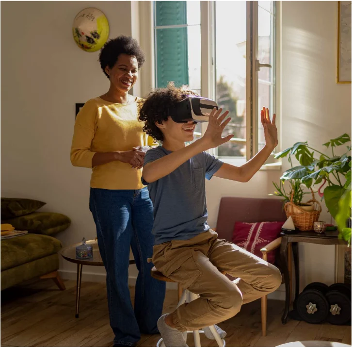 A boy on a stool reaches out while wearing a VR headset, a smiling woman in a mustard sweater behind him